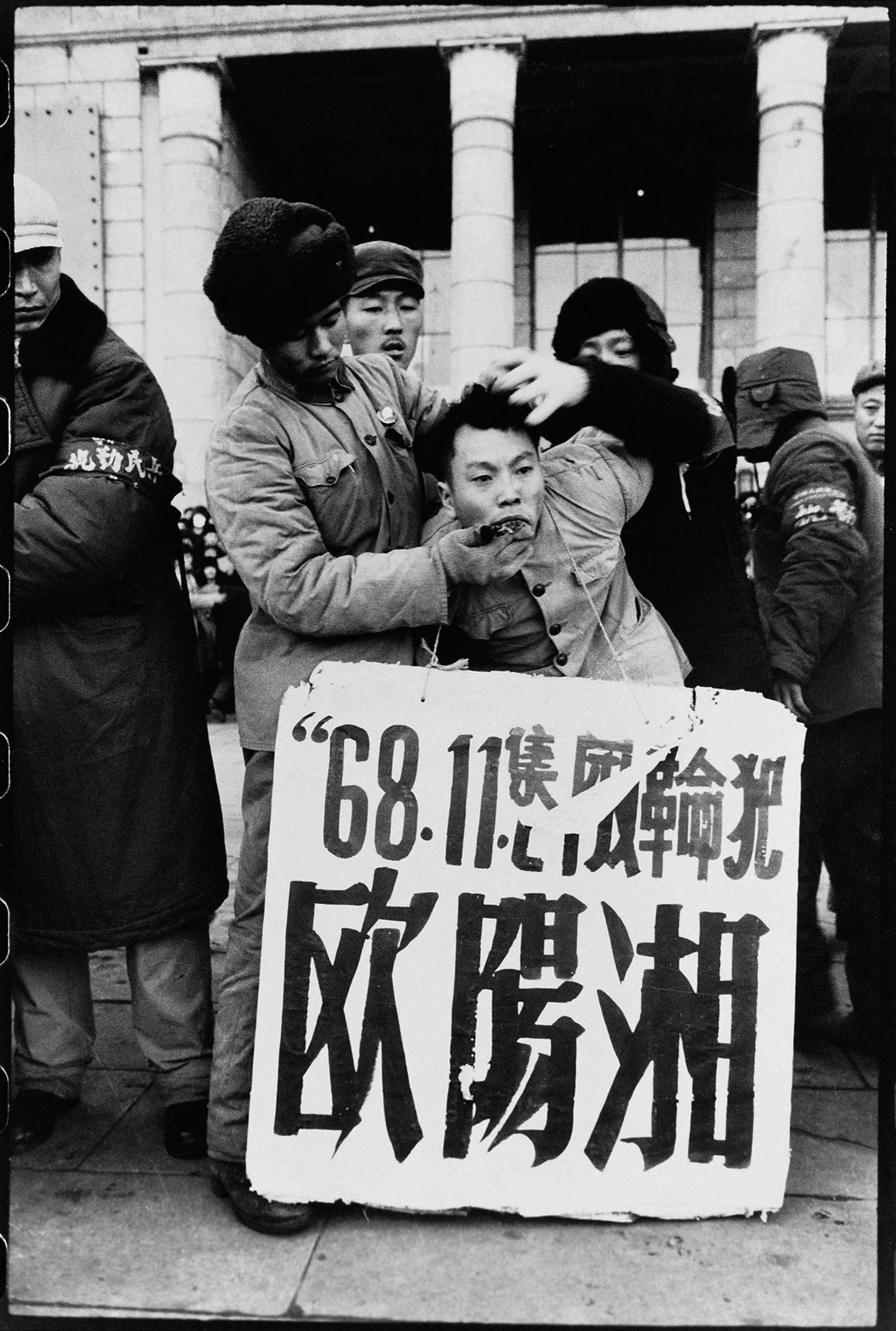 The son of a purged official endures a Struggle Session, the sign details the crime of the boy publically defending his father and the date of the offense.